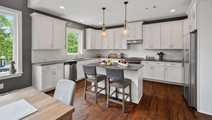 A modern kitchen with white cabinets, a central island with stools, pendant lighting, and a view of greenery outside. - Ruby Arrival by SoCharm | Luxury on Lake Norman (Mooresville)