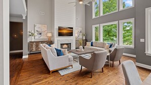 A spacious living room with a fireplace, large windows, and a ceiling fan. - Ruby Arrival by SoCharm | Luxury on Lake Norman (Mooresville)