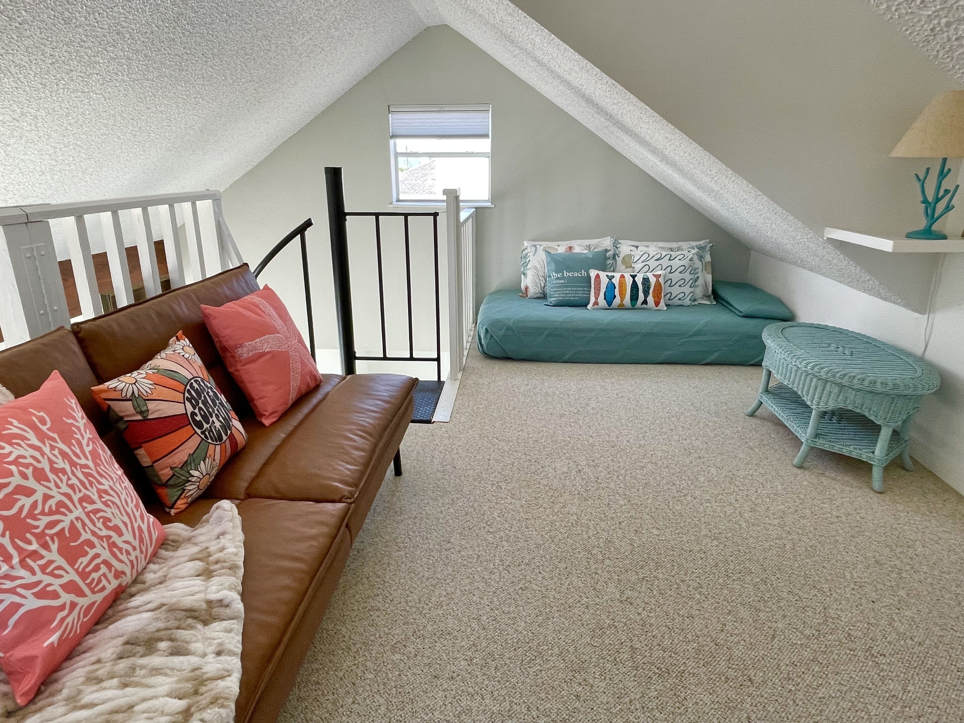 Een knus loft slaapkamer met een bruine leren bank, blauwgroene beddengoed en een klein rieten bijzettafeltje.