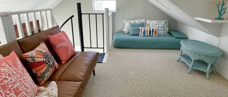 Een knus loft slaapkamer met een bruine leren bank, blauwgroene beddengoed en een klein rieten bijzettafeltje.