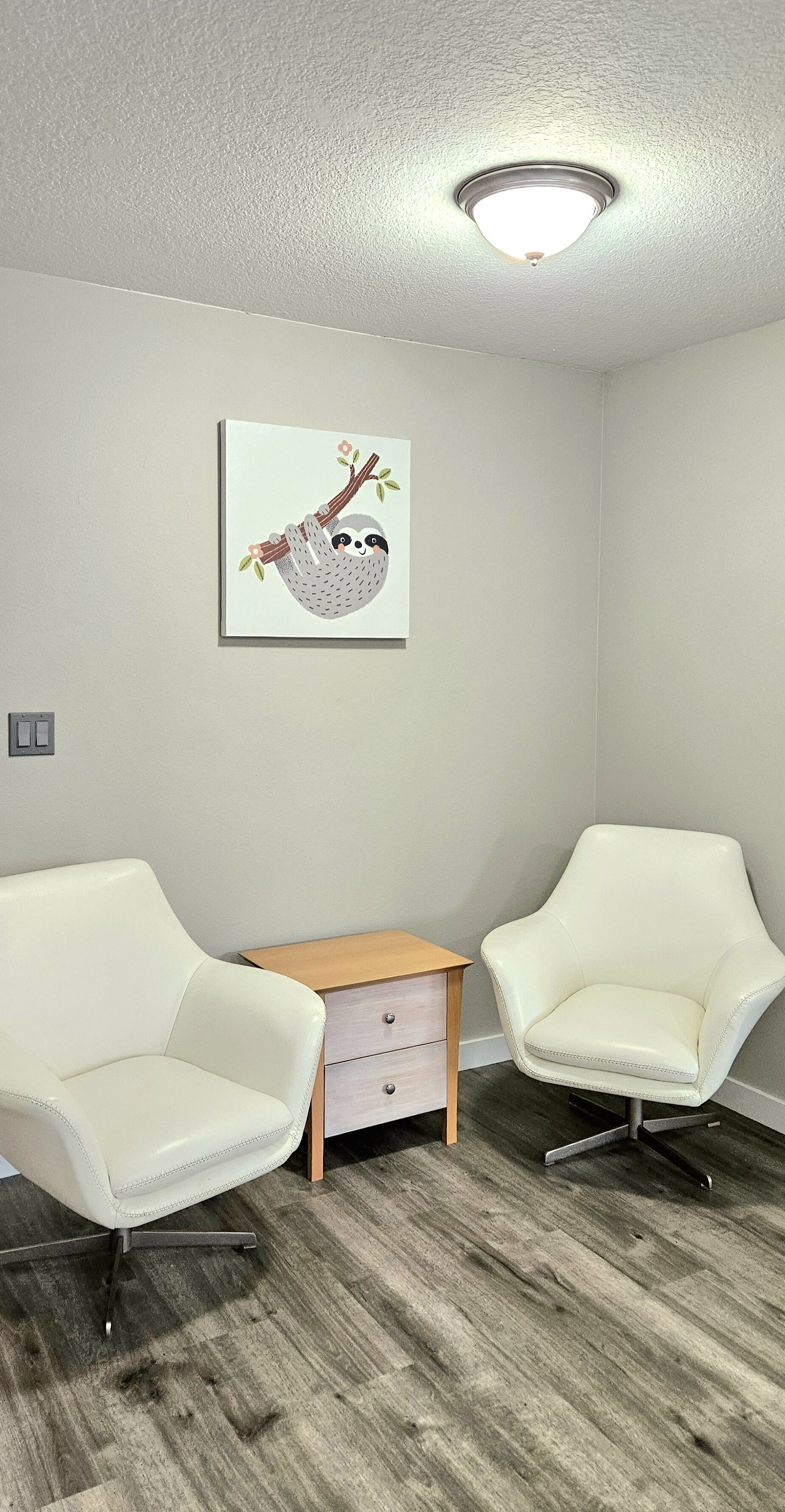 A room with two white leather chairs, a wooden side table, and a wall-mounted artwork of a sloth.