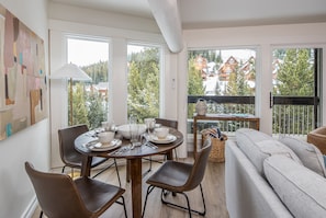 Un comedor moderno con una mesa redonda, sillas y un sofá. Amplias ventanas ofrecen vistas a una montaña nevada y edificios.