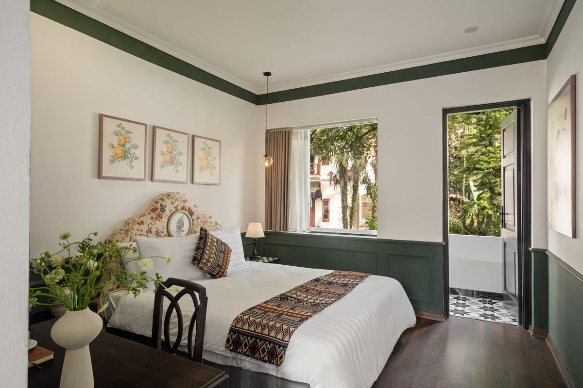 A neatly arranged bedroom with a large bed, a window with curtains, and framed botanical prints on the wall.