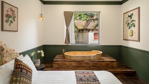 Une chambre avec une grande baignoire en bois, un lit agrémenté d’oreillers à motifs et une fenêtre donnant sur une maison.