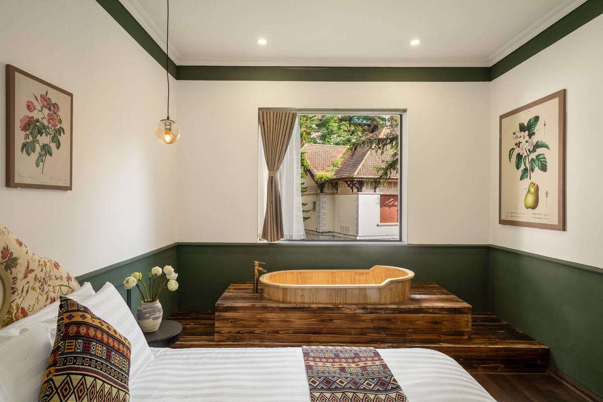 A bedroom with a large wooden bathtub, a bed with patterned pillows, and a window with a view of a house.