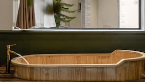 Une baignoire en bois se trouve dans une pièce dotée d’une fenêtre donnant sur une maison traditionnelle à toit de tuiles.