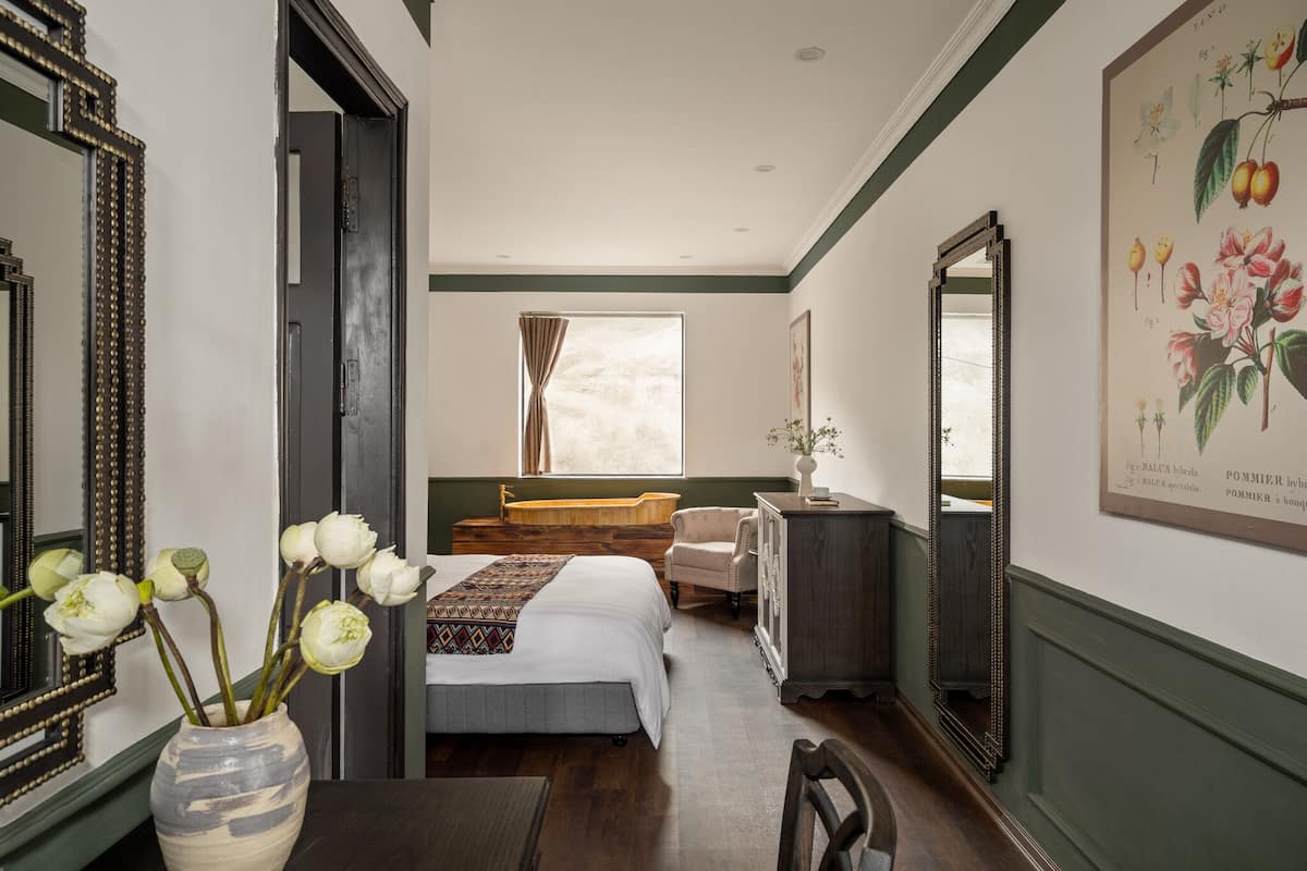 A bedroom with a bed, a chair, a dresser, a vase with flowers, and a large framed botanical print on the wall.