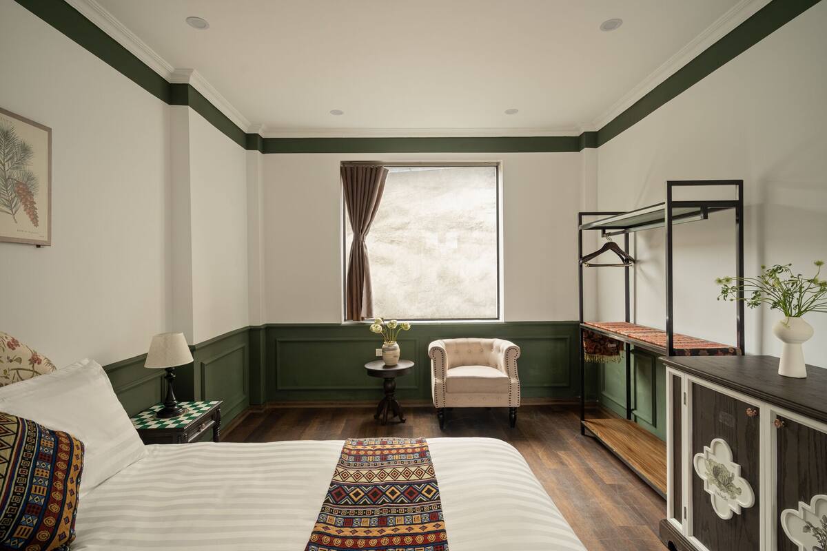 A hotel room with a large bed, a chair, a small table with a lamp, a window with curtains, and a dresser with a mirror.