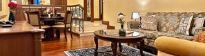Una sala de estar con una mesa de comedor de madera, sillas, un sofá con cojines estampados, una mesa de centro de vidrio y una escalera con barandilla.