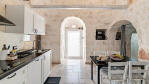 Una cuina amb armaris blancs, un taulell negre i una paret de pedra. Hi ha una zona de dinar amb taula i cadires, i una vista de l'exterior a través d'una porta d'arc.