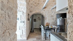 Interior de parets de pedra amb zona de cuina que inclou un frigorífic, una taula de dinar amb cadires i un sòl de rajola.