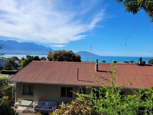 A house with a red roof, a small garden, and a view of mountains and a lake. - Sandy Feet Retreat - Enjoy a Classic Kiwi Batch with Ocean and Mountain Views (Kaikōura)