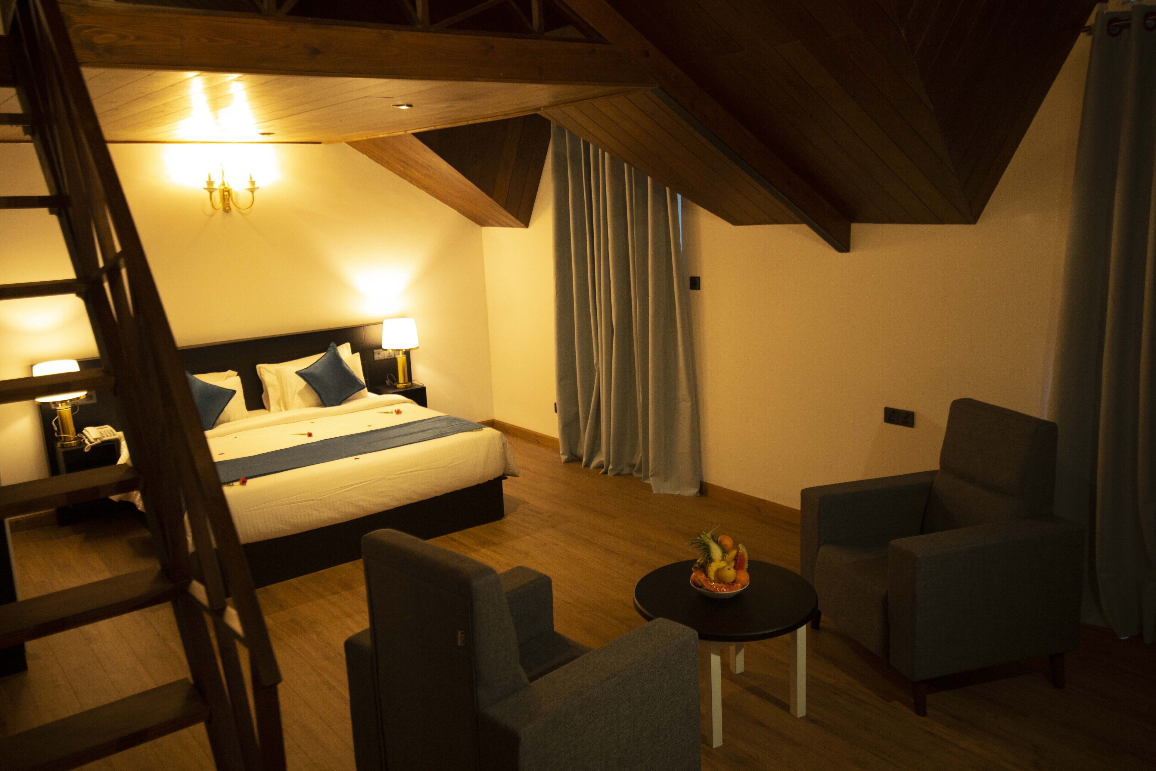 A hotel room with a bed, two armchairs, a small table with fruit, a chandelier, and a staircase.