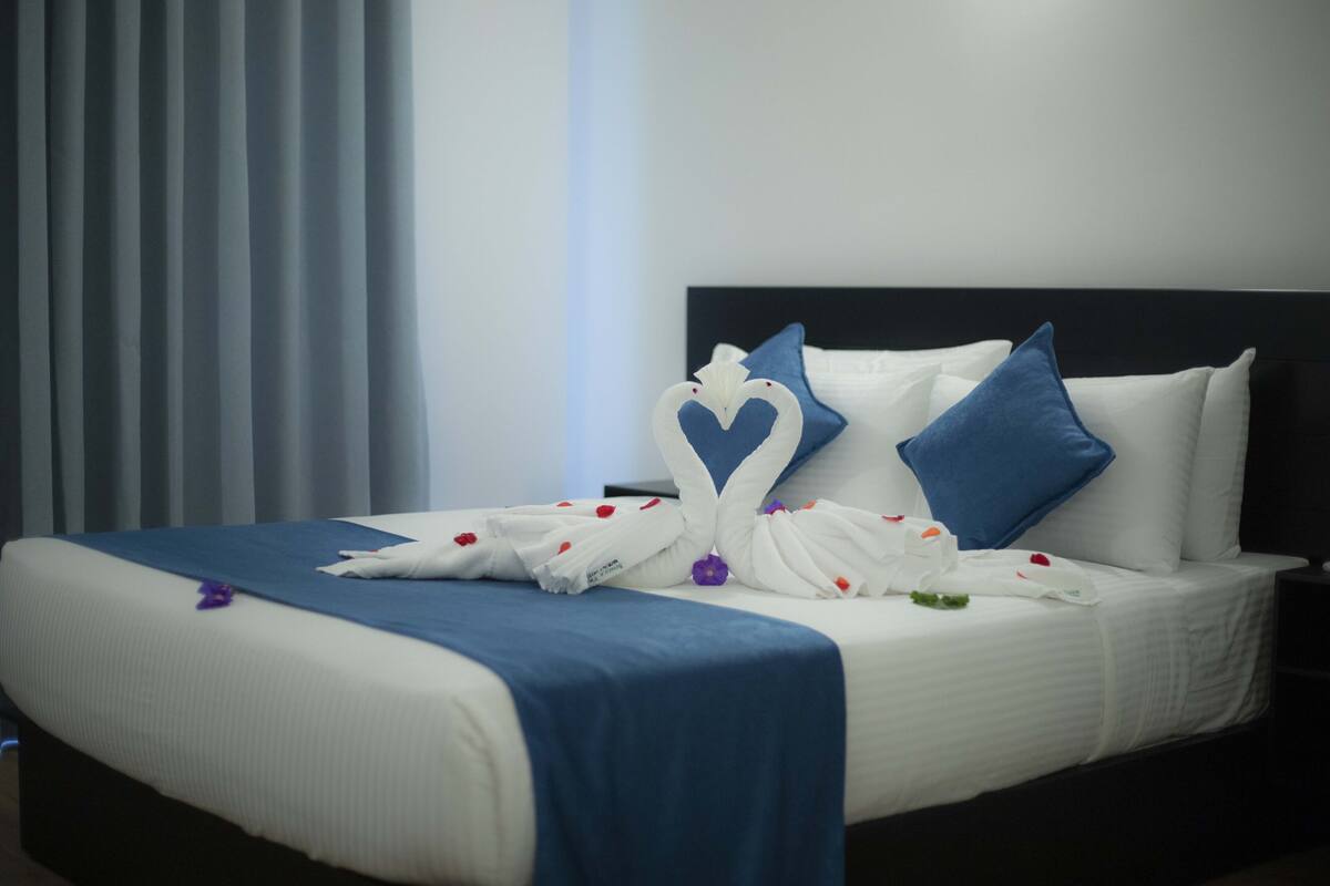 A neatly made bed with a blue and white bedspread, two blue pillows, and a heart-shaped towel decoration.