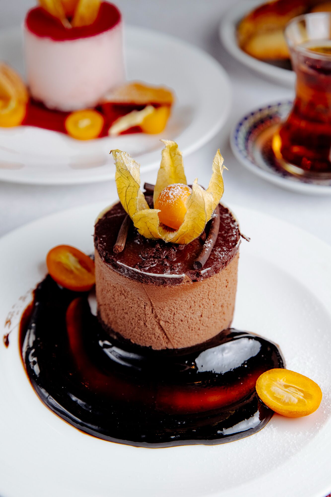 Un postre con mousse de chocolate, decorado con hojas amarillas y una cereza, sobre un plato blanco con salsa de chocolate negro.