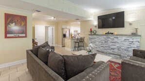 A modern living room with a grey sofa, a flat-screen TV mounted on the wall, and a kitchen in the background. - Tropical Vibes! Spacious 3BD/2BA, BBQ, Parking & more -10 min to Best Locations (West Palm Beach)