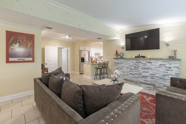 A modern living room with a grey sofa, a flat-screen TV mounted on the wall, and a kitchen in the background.