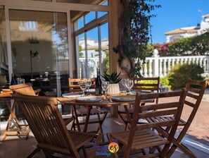 Un espacio al aire libre para comer con mesas y sillas de madera, una planta en maceta y vistas de edificios al fondo.
