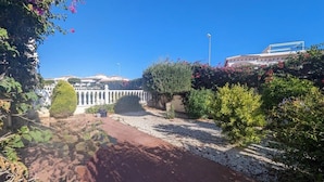 Un sendero de jardín que lleva a un edificio blanco con una fuente, rodeado de vegetación exuberante y plantas con flores.