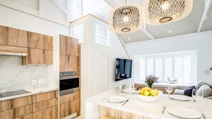 A modern kitchen with wooden cabinets, a built-in oven, and a large island with a bowl of fruit and wine glasses. - Seaside Escape Apartment Sea La View (Cape Town)