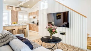 A modern kitchen with wooden cabinets, a dining area with a sofa and a small table, and a TV mounted on a white wall. - Seaside Escape Apartment Sea La View (Cape Town)