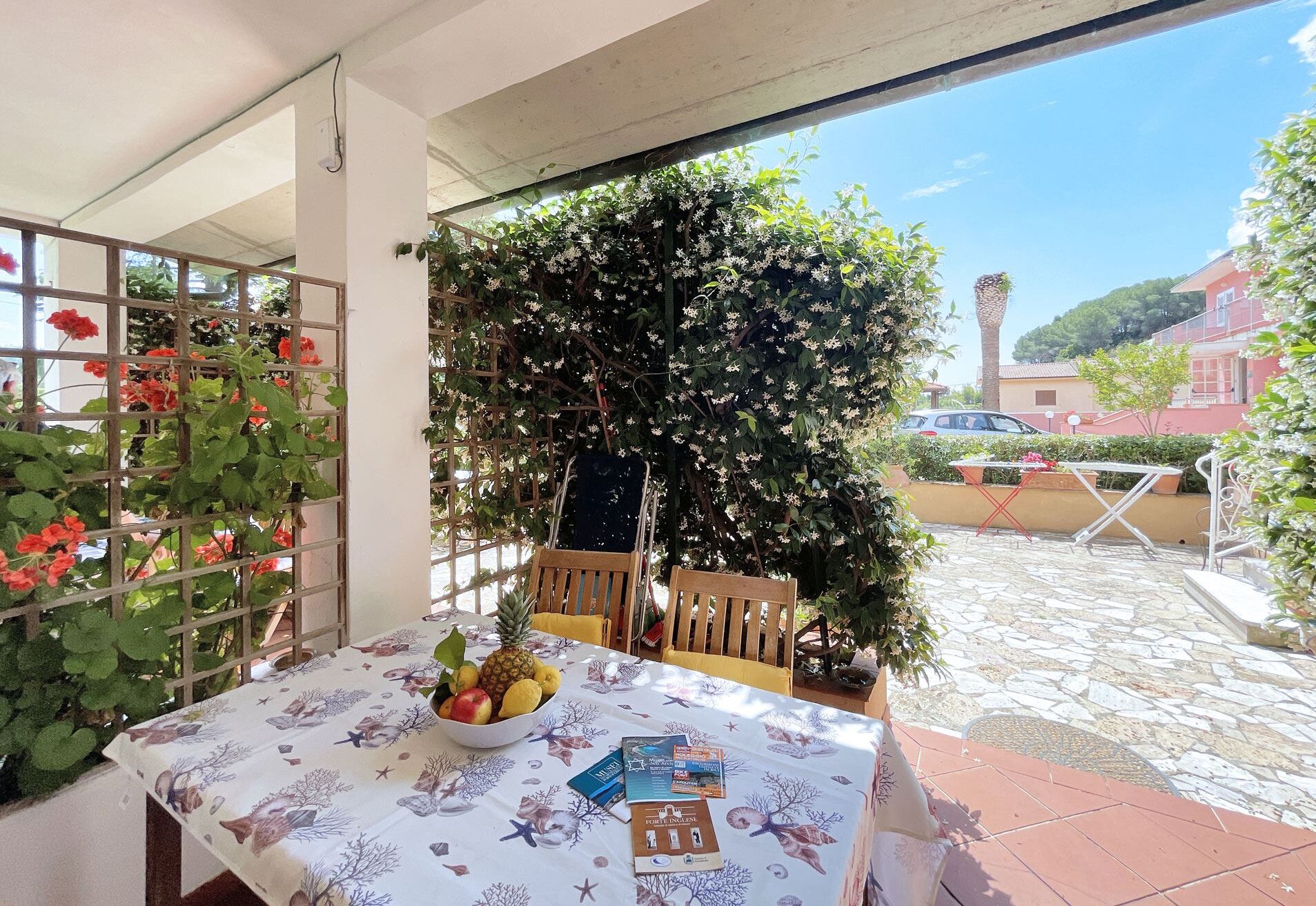 Une table avec un panier de fruits et des livres, une chaise en bois, et une vue sur un jardin avec des fleurs blanches et une voiture au loin.