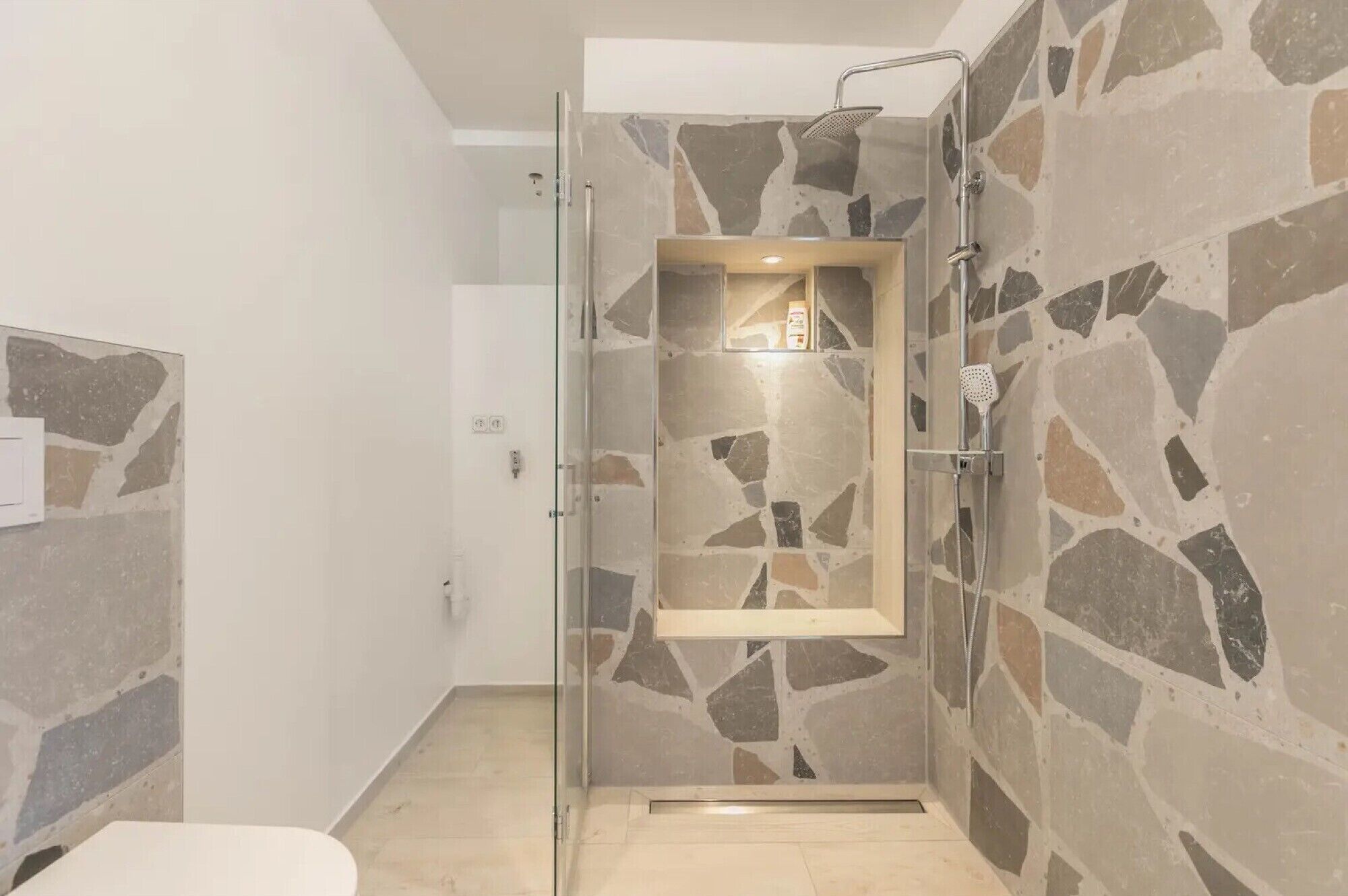 Una cabina doccia in vetro con parete in pietra, uno scaffale in legno e un water bianco.