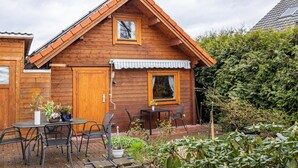 Puuinen mökki, jossa on harjakatto, pieni patio, jossa on tuoleja, pöytä ja ruukkukasveja.