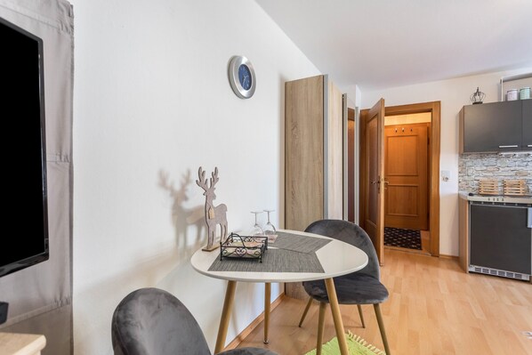 A compact living space with a round table, two chairs, a television, a clock, a wooden cabinet, and a kitchen area. - Studio Apartment 'Bergblick' with Mountain View, Balcony and Wi-Fi (Frasdorf)
