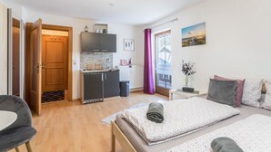 Quarto compacto com cama, pequeno espaço de cozinha, porta que dá para outro ambiente e janela com cortinas.