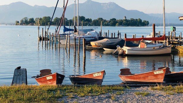 Um lago tranquilo com barcos atracados na margem, uma área gramada em primeiro plano e montanhas ao fundo.