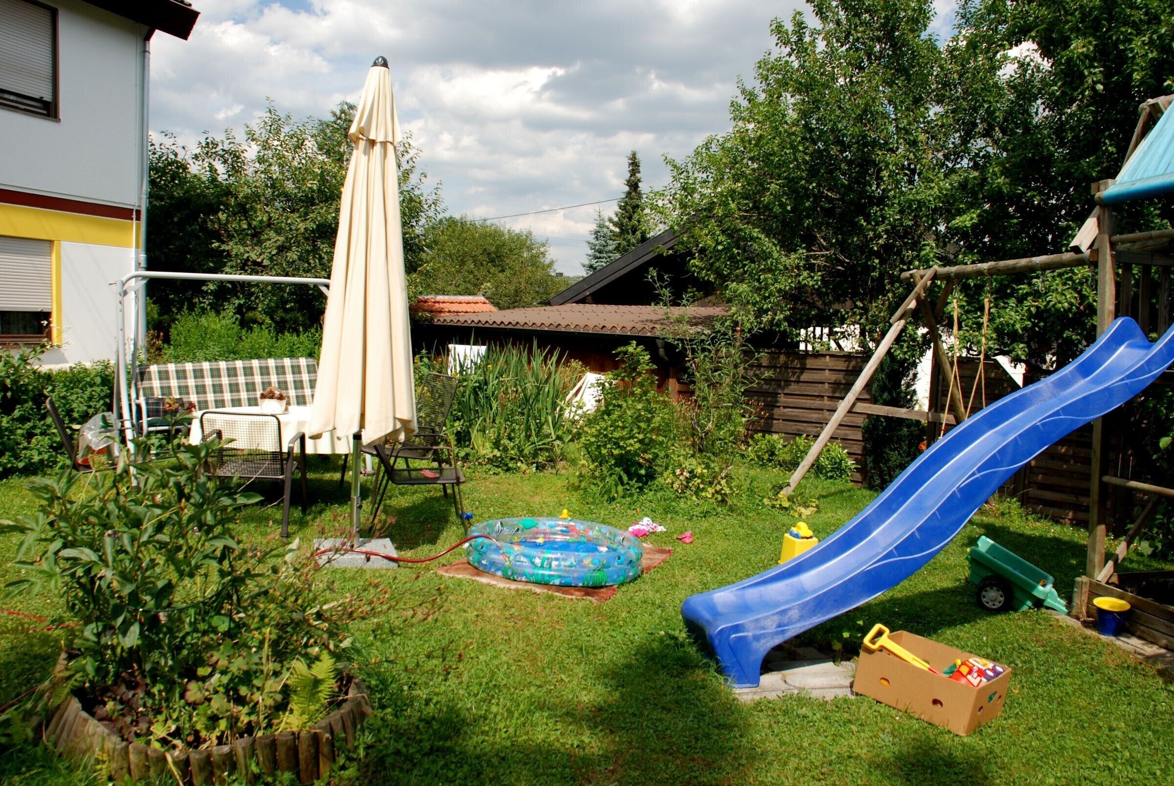 Patio trasero con tobogán, piscina inflable y casita de juegos de madera.