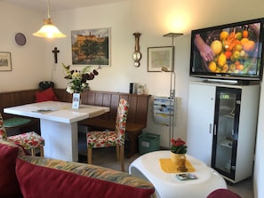 Une pièce comprenant une table à manger, des bancs, un téléviseur, un réfrigérateur et une horloge murale.