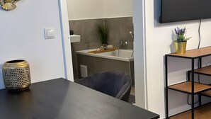Une chambre moderne avec une table noire, une chaise, un téléviseur, un miroir et une salle de bain visible à travers une porte ouverte.