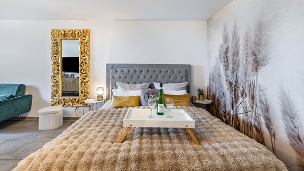 Une chambre à coucher avec une tête de lit capitonnée, un grand miroir doté d’un cadre orné, une table de chevet avec une lampe et une fresque murale.
