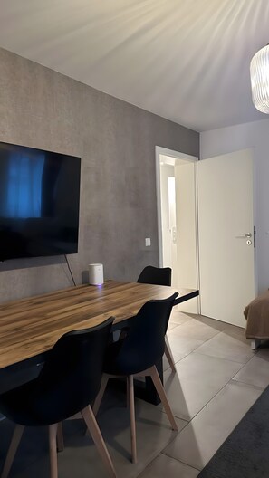 Un espace repas moderne avec une longue table en bois, des chaises noires, un téléviseur à écran plat fixé au mur et une porte blanche en arrière-plan.