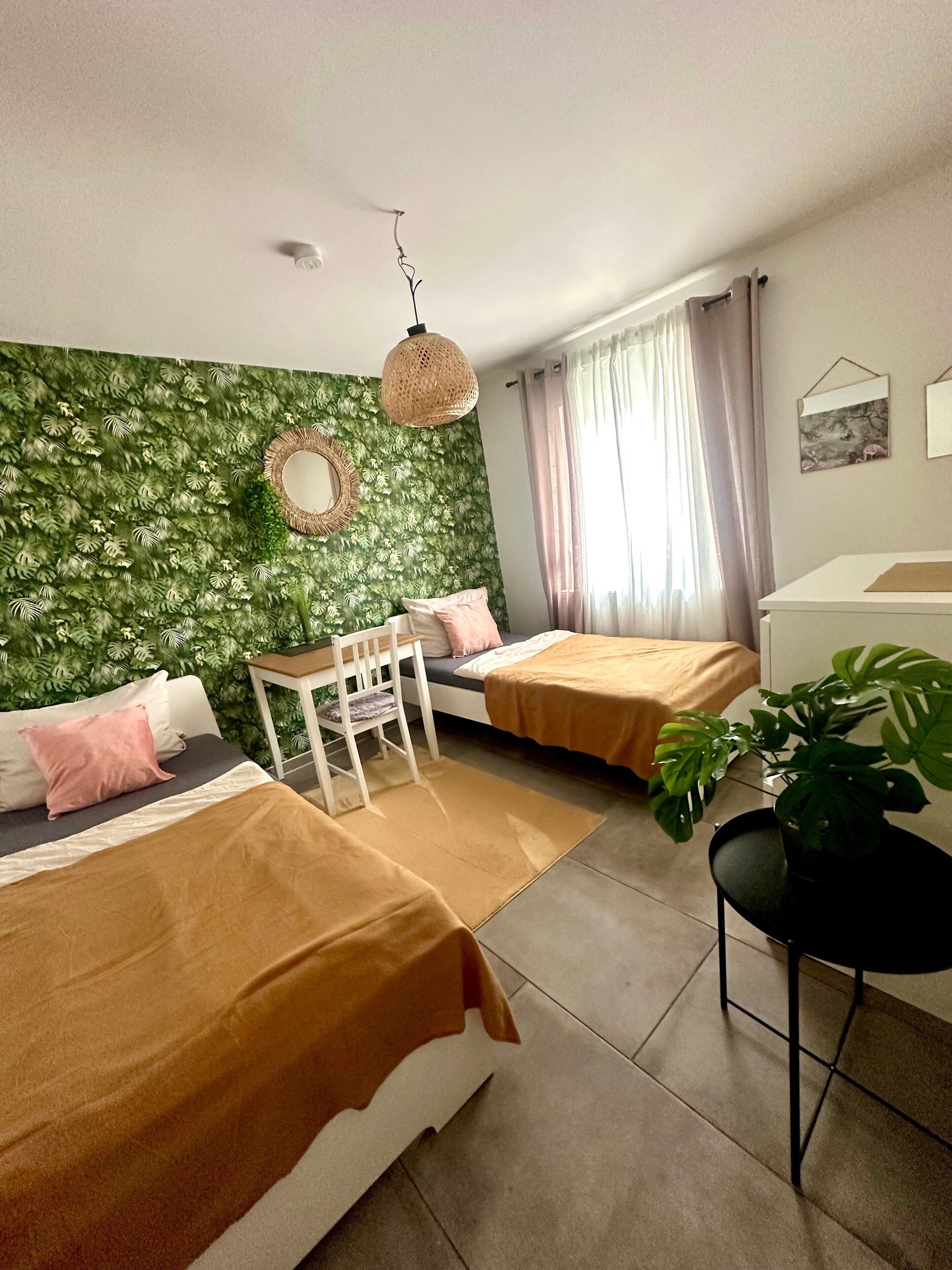 Ein Zimmer mit zwei Betten, einem kleinen Tisch, einem Stuhl, einer Pendelleuchte und einer Topfpflanze.