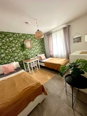 A room with two beds, a small table, a chair, a hanging light fixture, and a potted plant.