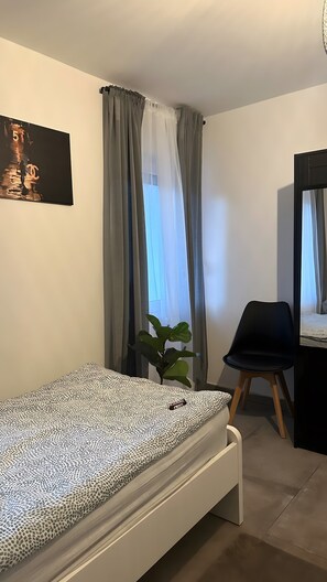 Une chambre à coucher avec un lit, une chaise, une plante, une fenêtre avec des rideaux et un tableau accroché au mur.
