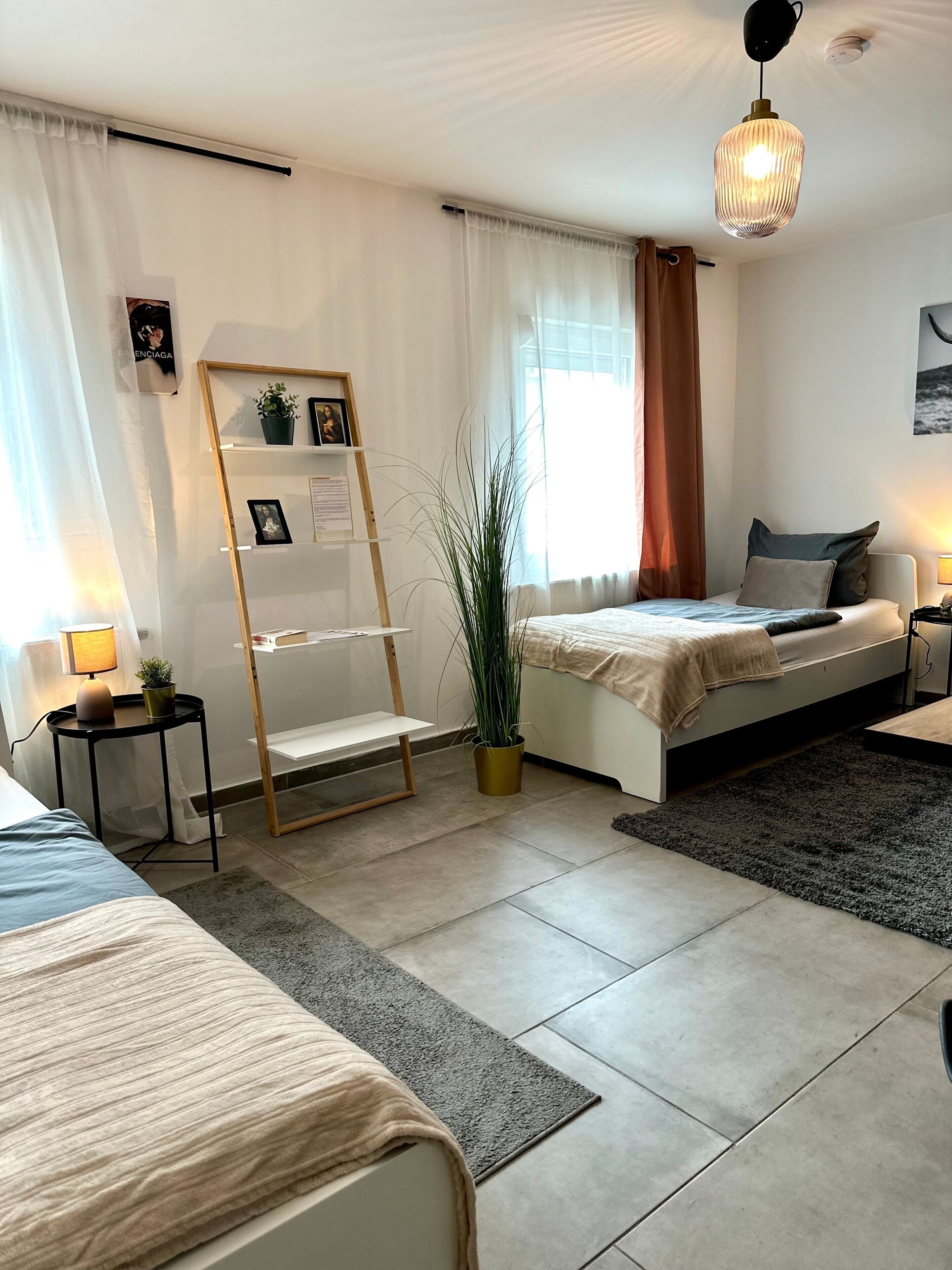 A modern bedroom with two beds, a bedside table, a floor lamp, a potted plant, and a window with curtains.