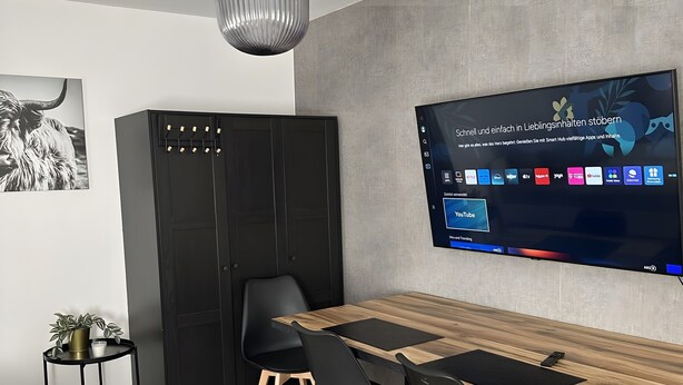 A modern dining area with a wooden table, black chairs, a black cabinet, and a mounted TV displaying a user interface. - Apartment 'Apartement Kingsize' with Shared Terrace and Wi-Fi (Duisburg)
