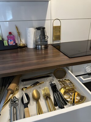 Une cuisine dotée d’un plan de travail en bois, d’une machine à café, d’une bouilloire, d’une râpe et d’un tiroir contenant divers ustensiles.