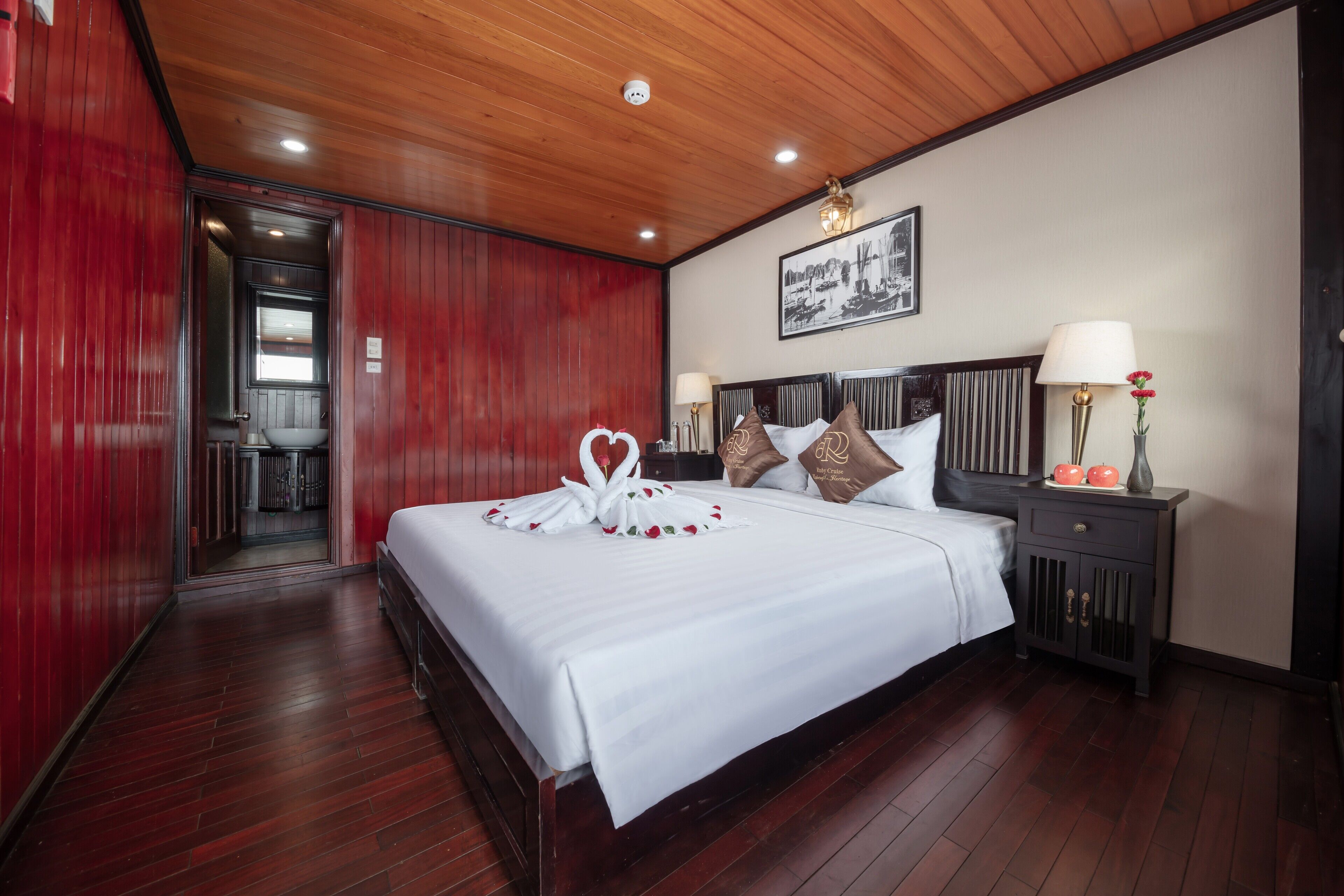 A hotel room with a wooden ceiling, a bed with white linens, a nightstand with a lamp, and a framed picture on the wall.