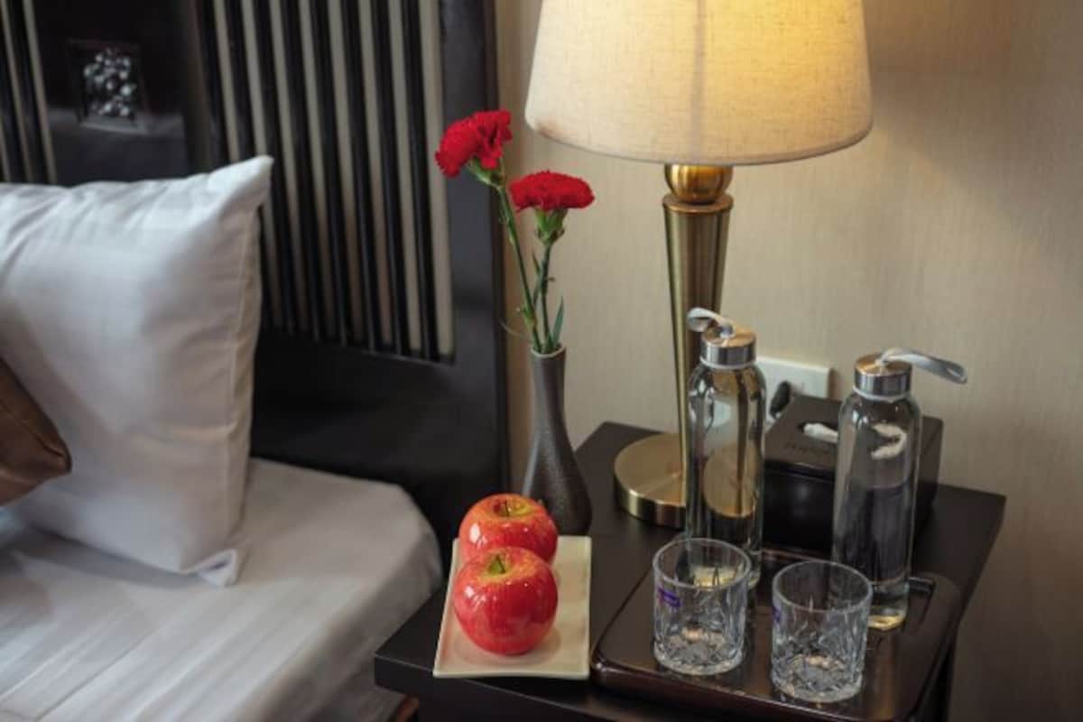 A neatly made hotel room with a bed, a bedside table with a lamp, two red apples, a vase with red flowers, and two glasses.