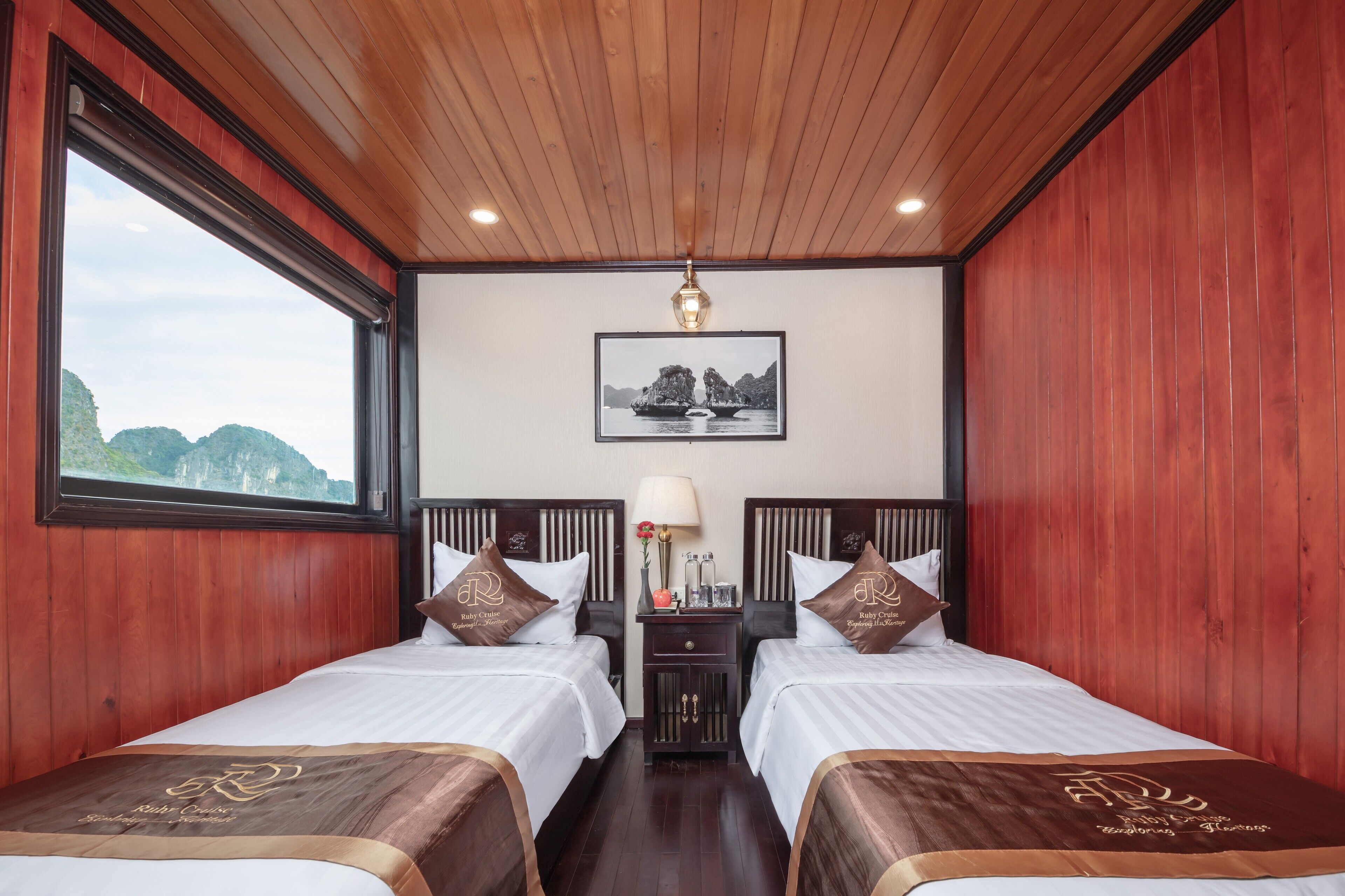Habitación de hotel con dos camas, paredes de madera, un ventanal grande, un cuadro enmarcado en la pared y una mesita de noche con lámpara.