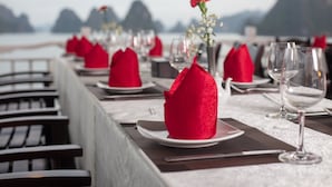 A dining table set with red napkins, white plates, and clear wine glasses, overlooking a body of water with distant mountains. - Ruby Cruise (Tuan Chau)