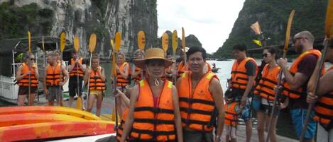 A group of people on a dock with life jackets and paddles, near a boat and rocky cliffs.