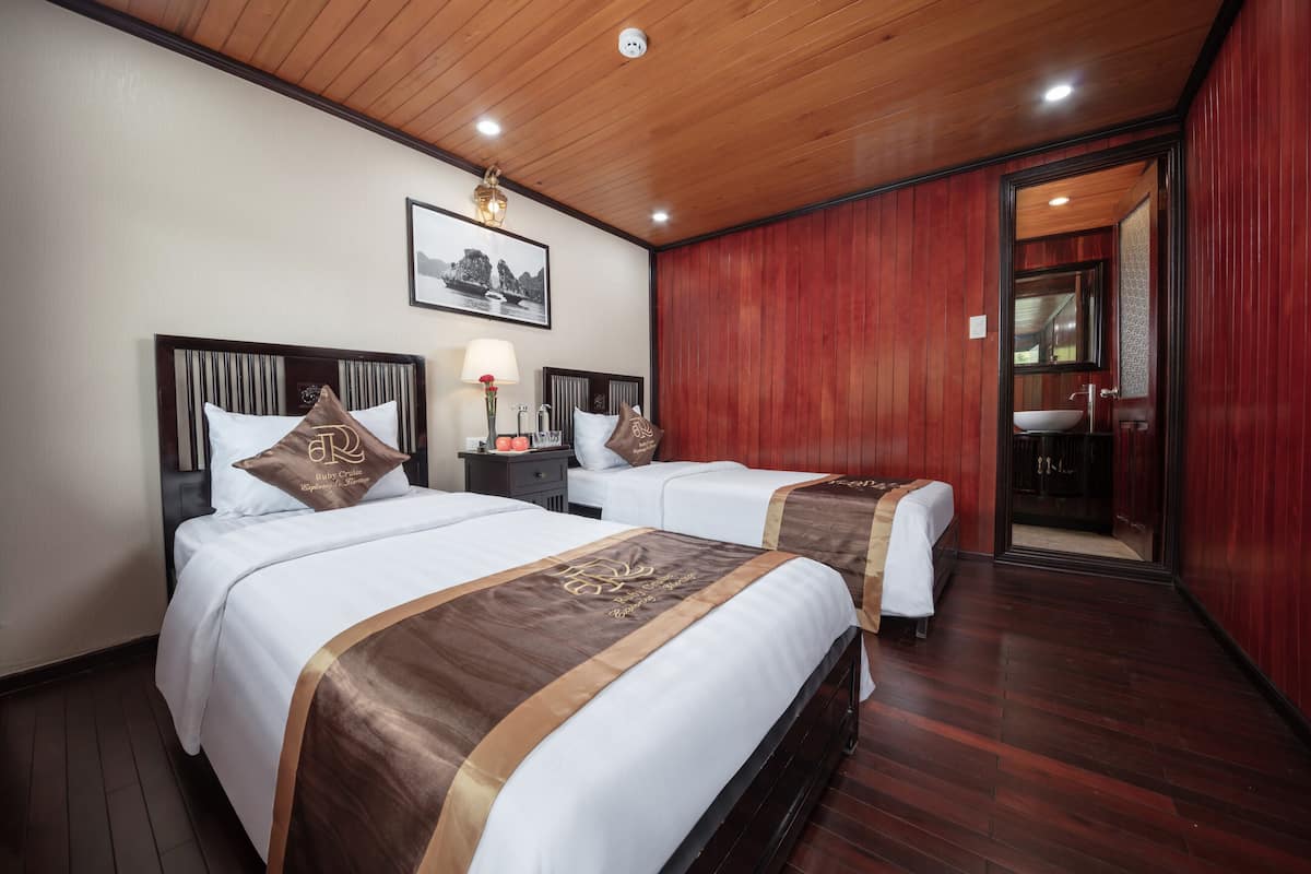 A hotel room with two beds, wooden flooring, a wooden ceiling, a framed picture on the wall, and a bathroom visible through an open door.