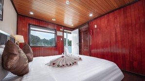 A hotel room with a wooden ceiling, a large bed with a white comforter and a towel arranged in a swan shape, a window with a view of greenery, and a door leading outside. - Ruby Cruise (Tuan Chau)