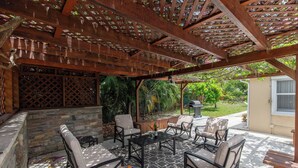 Un patio exterior con una pérgola de madera, pared de piedra y zona de estar con mesas y sillas.